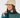 A woman wearing a cream ribbed knit pom-pom beanie with a small circular logo patch and a teal patterned knit golf sweater.
