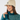 A woman wearing a cream ribbed knit pom-pom beanie with a small circular logo patch and a teal patterned knit golf sweater.