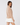 A woman wearing a beige mock-neck long-sleeve knit top and a white pleated mini skirt, shown from a side-back angle.