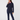 A woman modeling a dark navy blue two-piece tracksuit consisting of a long-sleeve quarter-zip fleece and matching pants, worn over a bright pink shirt, against a plain light gray background.