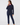 A woman modeling a dark navy blue two-piece tracksuit consisting of a long-sleeve quarter-zip fleece and matching pants, worn over a bright pink shirt, against a plain light gray background.