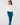 A woman wearing a white long-sleeve crewneck sweatshirt with a small black 'GOLF' with a star logo on the chest, paired with teal pants and teal socks.
