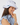 A woman wearing a white wide-brim sun hat with a black embroidered logo and a small blue-and-black strap detail, paired with a light blue ruffled blouse.