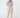 A woman stands barefoot against a light gray studio background, wearing a beige cap, a lavender and white patterned long-sleeve button-up shirt, and beige pants.