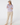 A woman stands barefoot against a light gray studio background, wearing a beige cap, a lavender and white patterned long-sleeve button-up shirt, and beige pants.