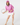 A woman modeling a pink long-sleeve half-zip top, white skirt, and pink cap, holding a tennis racket, front view.