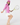 A woman posing in a pink quarter-zip top, white skirt, pink cap, and holding a yellow tennis racket, side view.
