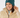 A smiling woman wearing a dark teal ribbed knit beanie with a small circular logo and a beige puffer jacket, photographed against a light gray studio background.