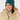 A smiling woman wearing a dark teal ribbed knit beanie with a small circular logo and a beige puffer jacket, photographed against a light gray studio background.