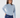 A woman wearing a light blue mock neck long-sleeve top and navy blue trousers, posing against a light gray studio background.