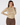 A smiling model wearing a long-sleeve mock neck top with a dense geometric pattern in beige, brown, and yellow, paired with brown pants, against a light gray studio background.